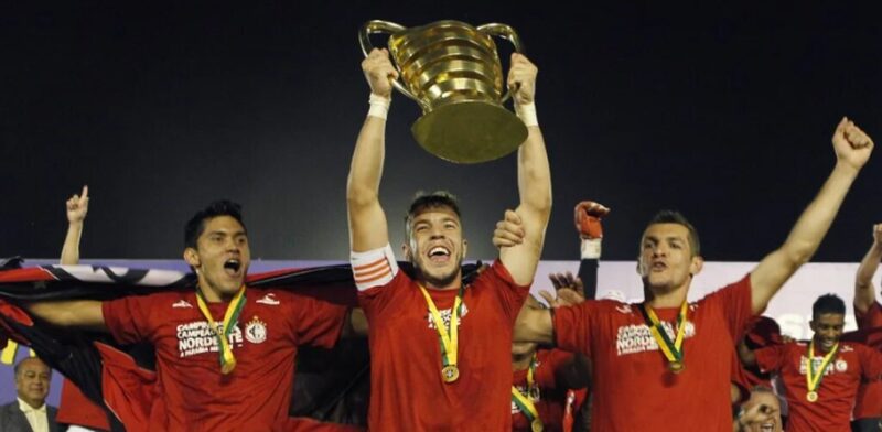 Capitão do Campinense levantando a taça do título da Copa do Nordeste em 2013, com outros jogadores com medalhas no pescoço celebrando ao redor do capitão