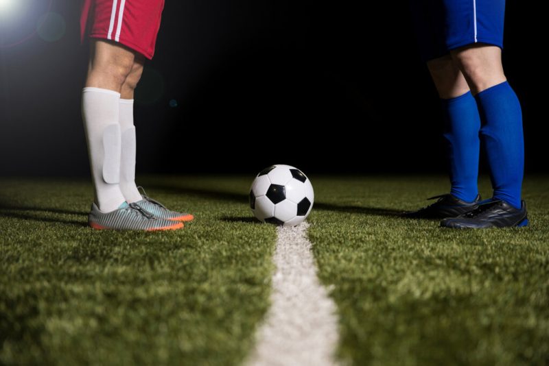 Dois jogadores rivais, frente a frente no meio de campo. Um com uniforme vermelho, o outro com uniforme azul. A bola está parada no centro do meio campi, simbolizando clássicos de futebol a Paraíba