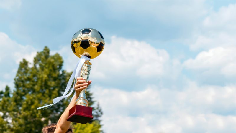 Simbolizando o time com mais títulos da Paraíba: Troféu de metal de bola de futebol sendo erguido ao ar livre, o céu está azul, claro e com nuvens. 